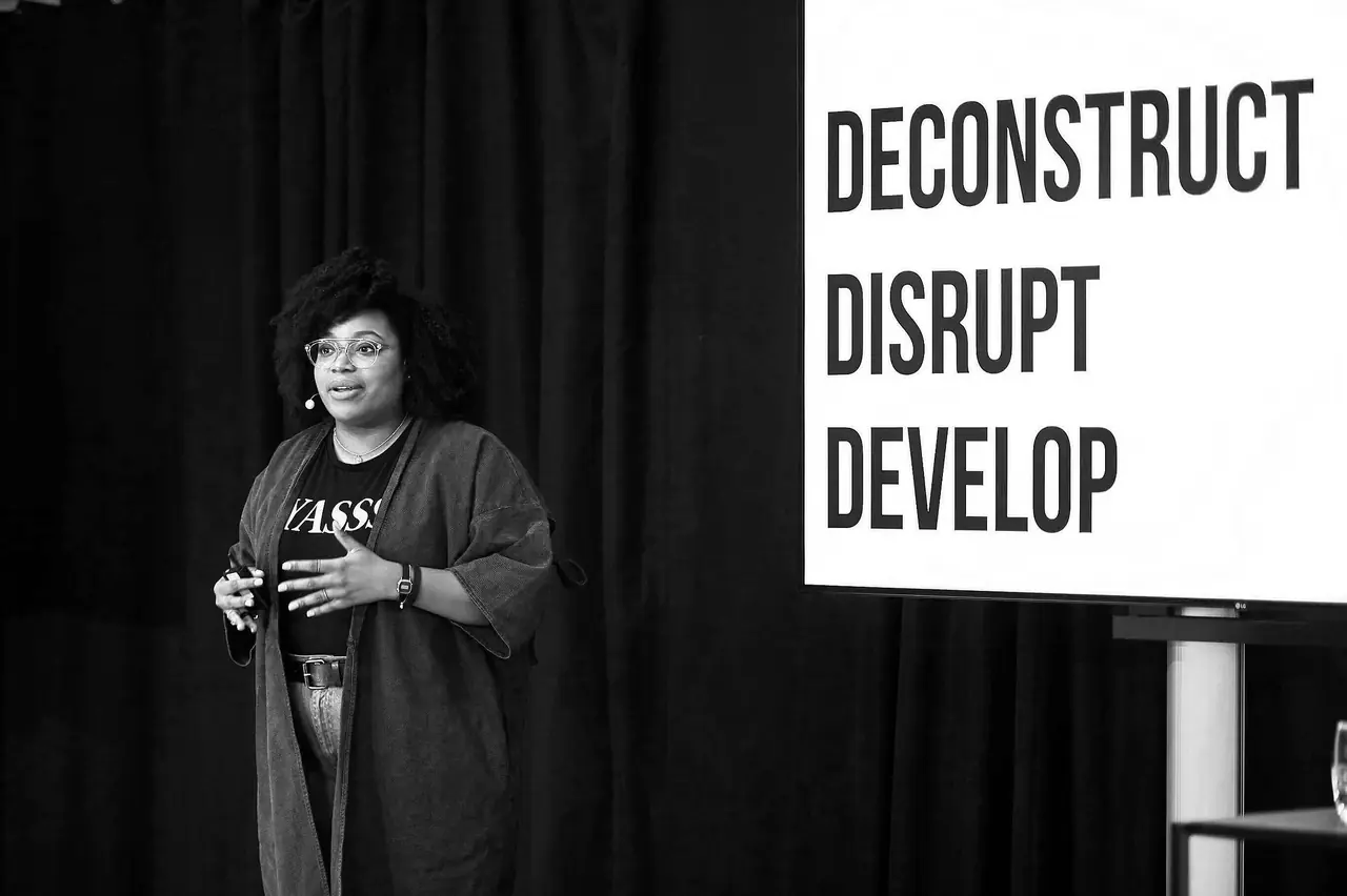 Vanessa speaking on stage beside a screen reading Deconstruct, Disrupt, Develop